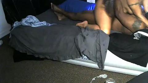 Media: Video of a dark-skinned man, shirtless, lying on a bed with a black sheet, holding a blue shirt. Background shows a white wall and a brown carpet.