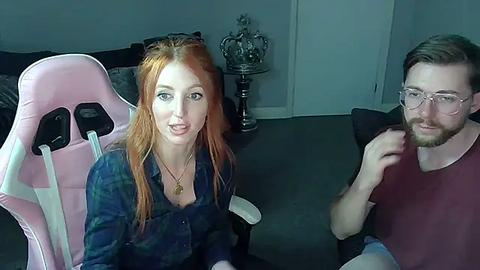 Media: A video of a red-haired woman in a blue shirt and a man with glasses and a beard, sitting on pink gaming chairs in a dimly lit, modern room.