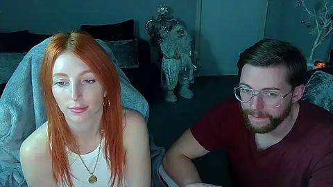 Media: Video of a fair-skinned woman with long red hair and a man with short brown hair and glasses, both wearing casual attire, sitting on a dark couch in a dimly lit room.