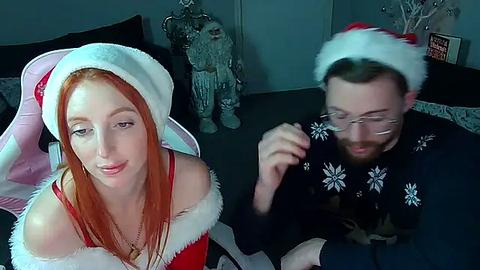 Media: Video of a fair-skinned woman with long red hair in a Santa hat and red dress, sitting beside a bearded man in a black sweater with reindeer pattern, in a dimly-lit room with Christmas decorations.