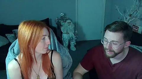 Media: Video of a young redhead woman with long hair in a black tank top, seated next to a bearded man in glasses, both smiling in a dimly lit room with blue walls.