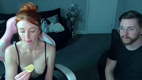 Media: Video of a red-haired woman in a low-cut black top, holding a chip, and a bearded man in glasses, both seated in a dimly lit room with a skull statue and a plush chair.