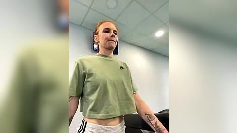 Media: Video of a fair-skinned, red-haired woman in a green crop top and white shorts, standing in a modern, white-walled room with recessed lights.