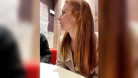 Media: Video of a young Caucasian woman with long, straight red hair, wearing a beige coat, seated at a red and white table in a modern, dimly lit restaurant with brick and glass walls.