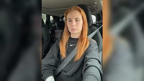 Media: Video of a young woman with long, straight red hair, wearing a dark gray sweatshirt, driving a car with a gray seatbelt, blurred background.