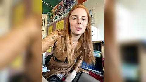 Media: Video of a young, fair-skinned woman with long, straight red hair, wearing a brown leather jacket, seated at a cluttered desk in a classroom.