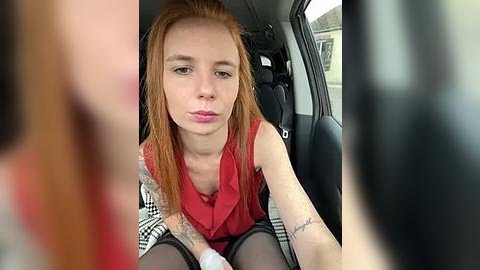 Media: A video of a pale-skinned, red-haired woman with a tattooed arm, pouting inside a car. She wears a sleeveless red top and checkered skirt. The background shows blurred people.