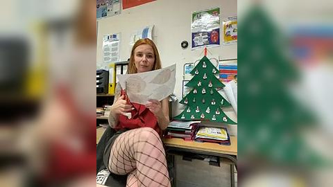 Media: A video of a red-haired woman sitting at a desk in a classroom, holding a paper. The background includes green Christmas tree decorations, bulletin boards, and a calendar.