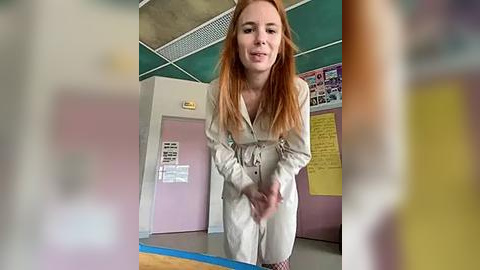 Media: Video of a young, fair-skinned woman with long, straight red hair, wearing a beige jumpsuit, standing in a classroom with green ceiling tiles, pink walls, and bulletin boards.