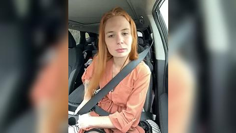 Media: A video of a young woman with long red hair wearing a beige shirt, seated in a car with a seatbelt on, blurry background.