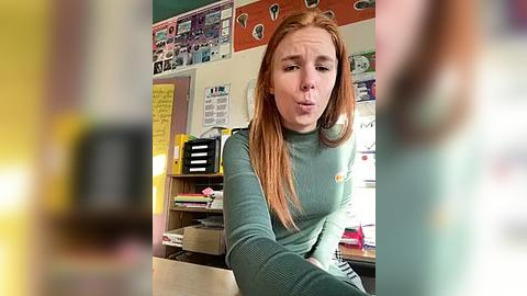 Media: Video of a young, fair-skinned woman with long red hair, wearing a green sweater, sitting in a cluttered classroom. The background features colorful posters and shelves with books and papers.