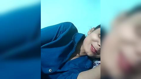 Media: A blurry, close-up video of a woman with fair skin, light brown hair, and red lipstick, wearing a blue blouse, lying on a bed with a blue background.