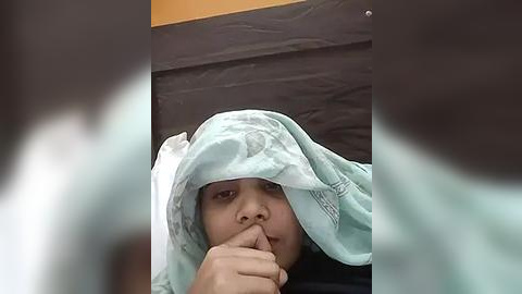 Media: Video of a young child with light brown skin, wearing a light blue blanket, hiding under it while covering their mouth with their fingers. Background shows a dark brown and beige cushion.