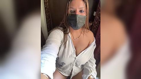 Media: Video of a young woman with light skin and long blonde hair, wearing a gray face mask and a white, unbuttoned shirt, revealing her chest. She's taking a selfie indoors with a blurred background.