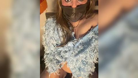 Media: Video of a woman with long brown hair, wearing a light blue, frilly top, black mask, and gauze over her eyes, sitting on a bed with a blurred background.