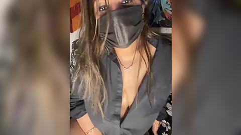 Media: Video of a woman with long brown hair, wearing a black face mask and unbuttoned black shirt, revealing cleavage. Background blurred, showing colorful patterns.