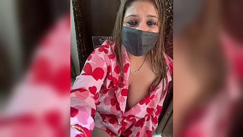 Media: A video shows a young woman with light skin and brown hair, wearing a red heart-patterned nightgown, grey face mask, and a visible cleavage. She sits on a wooden chair with a blurred red background.