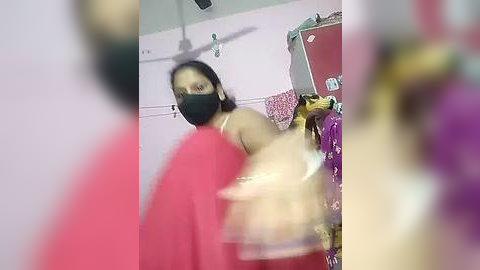 Media: Video of a woman in a pink saree and black mask, with blurred figures in the foreground, standing in a brightly lit room with pink walls and a fan.