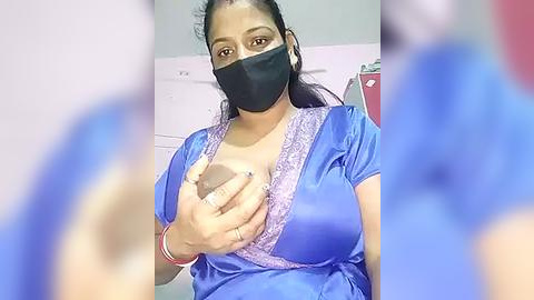 Media: Video of a woman with dark skin, wearing a blue satin robe with lace trim, black mask, and gloves, breastfeeding indoors.