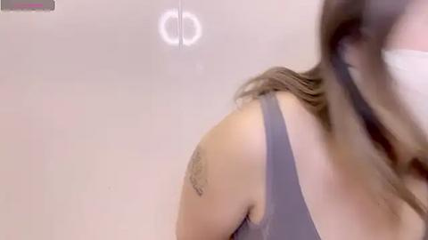 Media: A video of a woman with light skin, wearing a gray tank top, revealing a tattoo on her upper arm. Her hair is brown and partially obscured. The background is a plain, white wall with a circular light fixture.