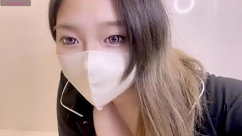 Media: Video of an Asian woman with long, dark hair, wearing a white face mask, black jacket, and light-colored top, indoors with a plain white background.