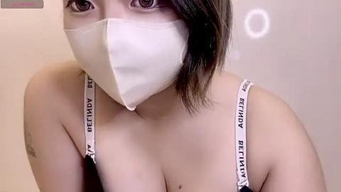 Media: Video of an Asian woman with medium-length black hair, wearing a white face mask, black bra straps, and a white top, against a blurred beige background.