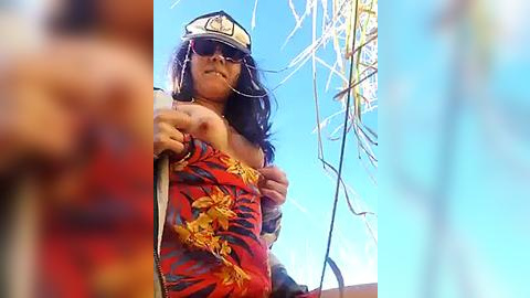 Media: Video of a tan-skinned woman with long dark hair and sunglasses, wearing a red and yellow floral top, standing on a boat under a clear blue sky.