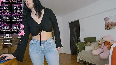 Media: Video of a young woman with medium-length wavy black hair, wearing a black crop top and blue jeans, standing in a cozy living room with a stuffed teddy bear and floral wall art.