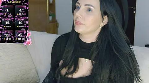 Media: A video of a fair-skinned woman with long black hair, wearing a black top, sitting on a beige sofa. The background shows a modern living room with a wooden cabinet and pink heart overlays.