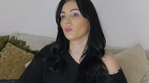 Media: Video of a light-skinned woman with long black hair, wearing a black off-shoulder top, seated on a beige couch with green and gold-patterned pillows in a neutral indoor setting.