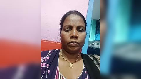 Media: Video of a middle-aged Black woman with medium skin tone, dark hair tied back, wearing a black floral-patterned blouse with a heart pendant necklace, against a pink and red wall.