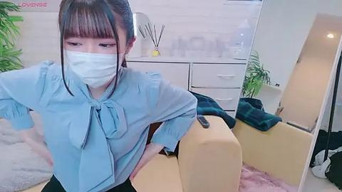 Media: Video of a young Asian woman with straight black hair, wearing a light blue shirt, mask, and sitting on a beige sofa in a modern, light-colored room with potted plants and a mirror.