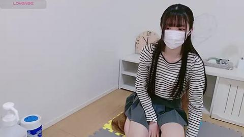 Media: Video of an Asian woman with long black hair, wearing a black and white striped shirt, grey skirt, and mask, sitting on a mat in a minimalist room with white walls, shelves, and a fan.