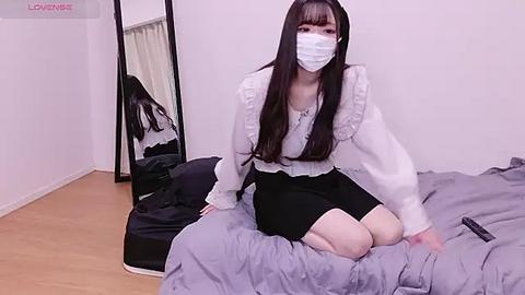 Media: Video of an East Asian woman with long black hair and pale skin, wearing a white frilled blouse, black skirt, and a face mask, kneeling on a grey bed in a minimalist, white-walled room.