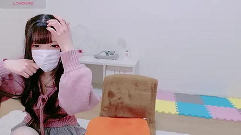 Media: Video of an Asian woman with long black hair, wearing a pink sweater and white mask, rubbing her head, seated in a room with a white desk, colorful puzzle mat, and beige chair.