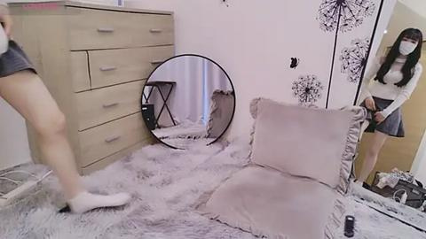 Media: Video of a bedroom with a beige dresser, a fluffy white rug, a round mirror, and a woman in a white sweater and gray skirt adjusting a cushion.