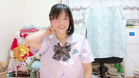 Media: Video of a smiling Asian woman in a white shirt with a plaid bow, standing in a laundry room with colorful clothes and a blue shirt on a drying rack.