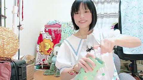 Media: Video of a young Asian woman with short black hair and light skin, wearing a white sailor-style dress, smiling and holding a green plastic bag in a cluttered, brightly lit room.