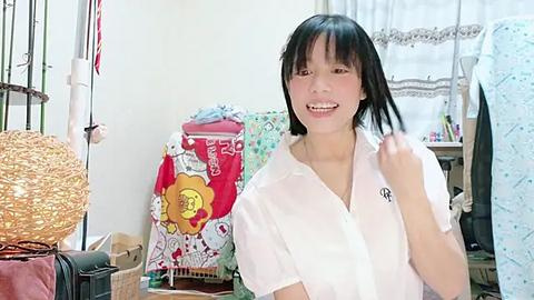 Media: Video of a smiling East Asian woman with straight black hair and fair skin, wearing a white shirt, standing amidst a cluttered room with colorful fabrics and a window with white lace curtains.