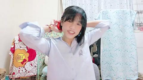 Media: Video of a young Asian woman with straight black hair, smiling, wearing a light blue shirt, holding a light blue pillow, surrounded by colorful, patterned pillows and a window with white curtains.