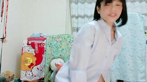 Media: Video of an Asian woman with short black hair, wearing a white lab coat, standing in a brightly lit room with colorful, patterned bed linens in the background.