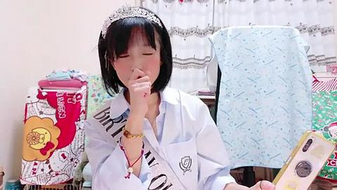 Media: Video of an Asian woman with straight black hair, wearing a white shirt with a sash, seated at a table with a smartphone. Background includes patterned fabric, boxes, and a phone case.