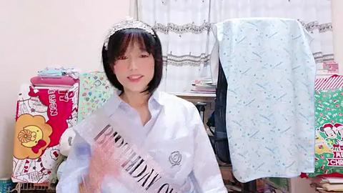 Media: Video of a young Asian woman in a white shirt with a sash, sitting in a cluttered room with colorful, patterned bedding and a computer.