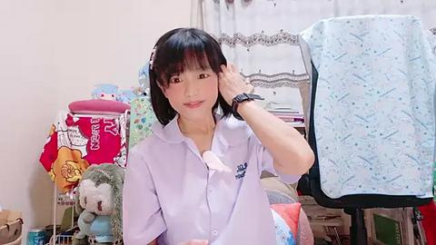 Media: A video of an Asian woman with straight black hair, wearing a light purple blouse, adjusting her hair in a cluttered bedroom.