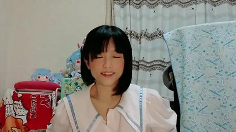 Media: Video of an Asian woman with short black hair, wearing a white sailor dress, smiling peacefully in a cluttered room with a red Hello Kitty container, a blue baby blanket, and lace curtains.