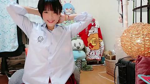 Media: A smiling young woman with short black hair, wearing a white button-up shirt and red skirt, sits on a chair in a cluttered, cozy room with plush toys, a wicker basket, and a decorative globe.