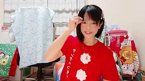 Media: Video of a smiling Asian woman with short black hair, wearing a red T-shirt with white flowers, adjusting a blue-patterned cloth on a mannequin in a cluttered bedroom.