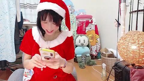 Media: A video of a young Asian woman in a red Santa outfit, with a festive room filled with plush toys, a Christmas tree, and a glowing lamp.