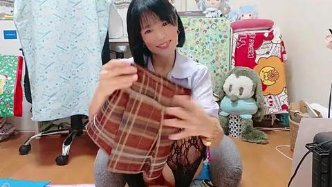 Media: Video of an Asian woman with black hair, kneeling on a wooden floor, holding a brown plaid shirt, smiling, surrounded by colorful fabric, stuffed toys, and a sewing machine in a cluttered room.