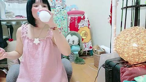 Media: Video of an Asian woman with black hair and pale skin, wearing a pink sleeveless top, drinking from a white baby bottle in a cluttered, brightly decorated room with stuffed animals, a red chair, and a wicker basket.